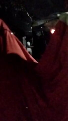 A nighttime outdoor scene shows several people gathered around a large, dark, and red-colored draped object. The object appears to be a costume or effigy, with parts of it moving. Some individuals are wearing traditional clothing, including head coverings. Lights are visible in the background, suggesting the presence of buildings or structures. The overall atmosphere is one of a public gathering or celebration.