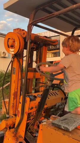 A young boy is playing on an old, orange forklift. The boy is wearing a grey t-shirt and green shorts. He's kneeling on a piece of wood placed on the driver's seat, holding the steering wheel with both hands. The forklift is rusty and appears to be out of service, parked in what looks like a yard with some greenery and other vehicles in the background. The sky is visible, illuminated with the warm light of either sunrise or sunset.