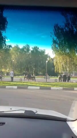 The video captures a scene from inside a vehicle looking out through the windshield. The view is of a park or outdoor recreational area with trees, a paved path, and benches. Several people are visible: one person stands near the path, two individuals sit on a bench, another person is on a bicycle, and a couple of other figures are seated on benches further down the path. The sun is shining, casting dappled light through the trees, and the sky is clear and blue. The overall impression is of a calm, pleasant day in a public outdoor space.