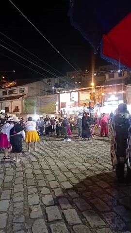 A night-time scene shows a cultural dance taking place on a cobblestone square, with spectators in the background. Dancers are dressed in traditional clothing, including hats, skirts and blouses. Some dancers wear masks and costumes resembling animal characters. The square is illuminated by overhead street lights, and buildings flank the square's edges. A flag partly visible in the foreground.