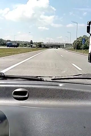 The video shows a first-person view from inside a car driving on a highway. The dashboard is visible in the foreground. The road is paved and has white lane markings. There are other vehicles on the road, including trucks and cars. In the distance, there is an overpass and trees on either side of the highway. The sky is blue with some clouds. The sun is shining.
