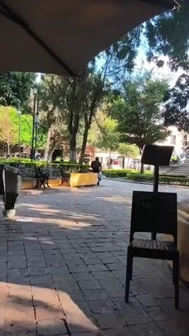 The video shows a public square, possibly in Mexico. The camera is positioned under a beige umbrella, looking out over a paved area made of square stones. A black chair is prominently placed in the foreground. Beyond the paved area is a green space with trimmed hedges and trees, along with several park benches. In the distance, a person wearing a black shirt is riding a scooter. In the background are buildings that suggest the location may be a town square. The light and shadows indicate that the video was filmed during daylight hours.