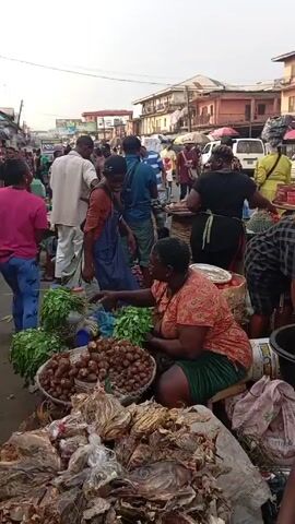 The video captures a busy street scene in what appears to be a market or commercial area. Various people are present, including vendors selling goods such as tomatoes, dried fish, and bananas. Some individuals are walking through the crowd, while others are riding in a three-wheeled vehicle called a "keke." Buildings are visible in the background, with signs and awnings lining the street. The overall atmosphere suggests a bustling and active urban environment.