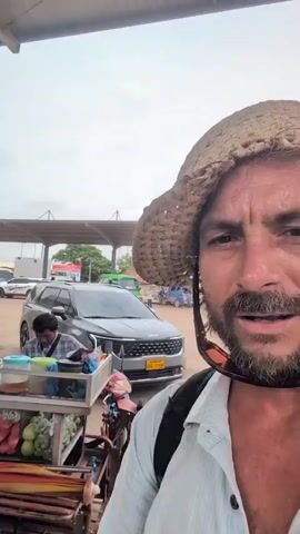 The video begins with a close-up shot of a man's face, who is wearing a straw hat and sunglasses pushed up on the hat. He is looking directly at the camera with a slightly concerned expression. He is wearing a light-colored collared shirt and a backpack. The background is slightly out of focus, but it appears to be an outdoor setting with vehicles and a vendor stall.

The camera then pans to the left, revealing more of the scene. A red scooter with a sidecar is parked in front of a food stall. The stall is stocked with various fruits and vegetables, including bananas, apples, and what appear to be gourds. A person is seen behind the stall, interacting with the items. Several vehicles are parked in the background, including a gray minivan and a white bus. The environment looks like a transportation hub or a market area, possibly a bus station or a roadside stop. The sky is overcast. The man in the foreground is still visible in the frame, though less prominent. The video appears to be a selfie-style recording by the man.