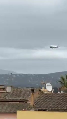 The video shows a white and yellow airplane approaching for landing. The plane is flying in front of a background of rolling hills and mountains on a cloudy day. In the foreground are red-tiled roofs of buildings, with a white satellite dish visible on one. The plane gradually descends closer to the ground as it approaches.