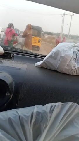 From the perspective of a car interior, the video shows a dusty roadside scene. Several people, mostly women, can be seen. Some wear hijabs or other modest clothing. A yellow auto rickshaw is visible in the background. In the foreground, on the car's dashboard, there are two white plastic bags. The background appears dusty or hazy.