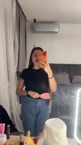 A woman is taking a selfie in a bedroom mirror. She is wearing a black t-shirt and blue jeans. In her left hand, she holds a pink hairbrush. She purses her lips into a kissy face for the camera. A child can be seen standing behind her to the left. The bedroom contains a bed with a grey headboard, beige pillows, and a patterned comforter. There is also an air conditioner unit on the wall. A vanity is in front of the mirror with a ring light and miscellaneous items on it. Curtains hang to the left of the mirror.