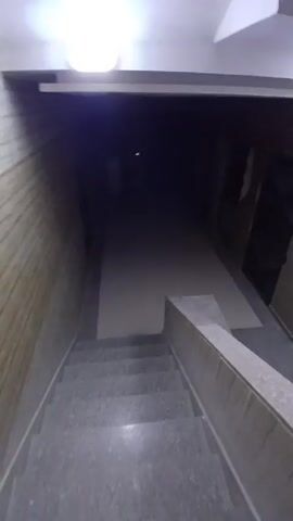 The video presents a first-person perspective from the top of a staircase leading down to a dimly lit hallway. The hallway has a smooth, light-colored floor. The walls are a mix of textured material and what appears to be exposed brick, with doors or openings along the right side. The ceiling is low and modern, with rectangular recessed lighting. The overall impression is of an unfinished or abandoned space, with a stark and somewhat unsettling atmosphere. The lighting is poor, further obscuring details and emphasizing the darkness at the far end of the hallway.