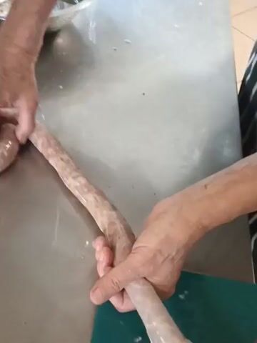 A person's hands are shown stuffing sausage into a casing. The person is holding a long casing and pushing it onto a stuffing tube. A coiled, partially stuffed sausage is visible to the left. The scene appears to be in a kitchen or food preparation area, with a stainless steel countertop.