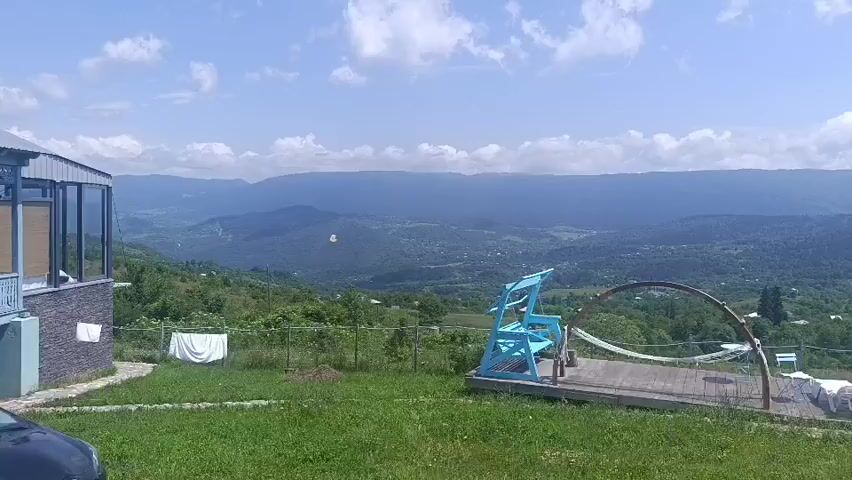 A video shows a traditional house with a blue decorated porch and a black car parked in front of it. The house is situated on a green hill with a valley and mountains in the background. A blue swing set is visible in the yard, and white laundry is hanging on a clothesline. The sky is partly cloudy. The video appears to have been filmed in a rural area.