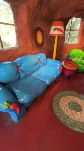 The video shows the inside of a room resembling a cartoon rabbit's dwelling. The walls are textured like earth or clay. The room is furnished with a bright blue, rounded sofa, a green, rounded armchair, a small round table, and a yellow lamp with a lampshade depicting a carrot. The floor is red, with a round green rug in the center. Light enters through two windows. There's also a framed picture on the wall. A trophy case labeled "TROPHIES" is visible set into the wall. People are walking around in the background.