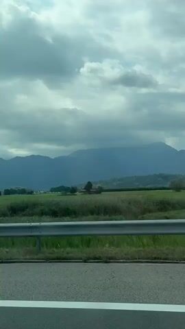 The video shows a view from a moving vehicle looking out over a green landscape with mountains in the distance. A metal guardrail runs across the foreground, separating the road from a grassy area. The sky is overcast with dramatic clouds. The landscape appears to be rural, with fields and trees visible. There are no people or animals explicitly shown. The footage seems to be captured during daylight hours, under what appears to be cloudy weather conditions.