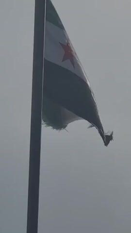 The video shows a tattered Syrian flag waving in the wind against a grey sky. The flag is mounted on a metal pole and appears worn, with frayed edges. The flag's design includes green, white, and black horizontal stripes with three red stars on the white stripe. The sky is overcast.