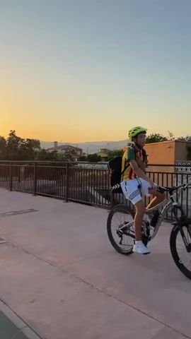 The video captures a picturesque sunset scene in Grao, Spain. The sun, a bright orb of warm orange and yellow, is setting behind a range of distant mountains, casting a golden glow across the sky. The sky itself transitions from a pale orange near the horizon to a soft, clear blue overhead.

The foreground features a paved walkway with a brown metal railing, suggesting a public area like a promenade or a bridge. On the walkway, a person is seen riding a bicycle, their silhouette visible against the bright sky, indicating they are moving away from the camera.

Further back, beyond the railing, there's a glimpse of a canal or water feature, with some greenery growing along its edges. Trees, one with flowing branches prominently positioned, dot the landscape, adding to the natural beauty. In the distance, buildings and other structures are visible, hinting at a residential or semi-urban setting. A streetlight with a distinctive modern design stands near the walkway.

The overall mood of the video is serene and peaceful, enhanced by the warm colors of the sunset and the tranquil atmosphere. The time of day is clearly sunset, and the weather appears to be clear and pleasant. There are no visible text elements within the video. The presence of the bicycle rider suggests casual leisure activity.