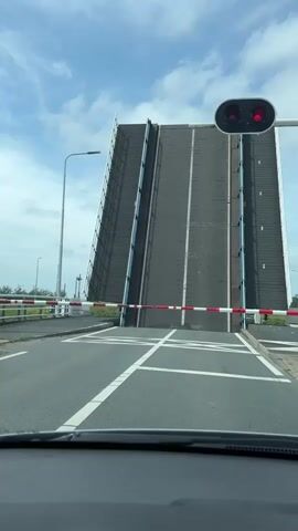 A driver's perspective captures a drawbridge opening, revealing an operator's control tower with a warning sign. The drawbridge is closed, with a red light, and two traffic barriers are down. The sky is partly cloudy.