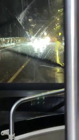 The video shows a view through the windshield of a vehicle, possibly a bus, at night during a rainstorm. Raindrops are visible on the windshield and streaks from the wipers can be seen. The road ahead is wet and reflective, with the headlights of oncoming cars creating bright glares and illuminating the rain. There are yellow and white lane markings on the road. A yellow barrier is visible on the left side of the road. In the reflection on the windshield, silhouetted figures of people are visible inside the vehicle. The environment appears to be an urban street.
