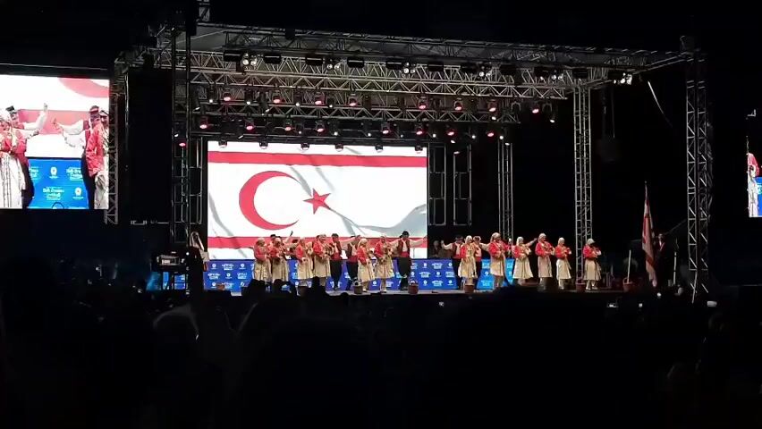 A group of approximately 20 performers in traditional attire are on a stage in front of a large screen displaying a flag. The performers are arranged in two rows, with a man in a darker outfit in the center of the front row, seemingly leading the group. They are all facing the audience and appear to be in the middle of a performance, possibly dancing or singing. The stage is lit by numerous stage lights. The screen behind them shows a white flag with a red crescent moon and a red star. To the right of the stage, a flag on a pole is visible. In the foreground, the silhouettes of an audience are visible. The overall environment appears to be a large indoor venue.