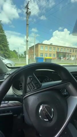 The video provides a first-person view from inside a car, looking out through the windshield and side windows. The **scene** is an outdoor setting, likely a residential or community area, with a **building** that appears to be a school or similar institution in the background. The building is light brown with multiple windows and is situated behind a fence. In front of the building, there are two large trash bins, one blue and one yellow.

Several **objects** are visible: the car's steering wheel, dashboard with a digital display showing "27°C" and a warning icon, the car's infotainment system displaying "Bluetooth Audio," a rearview mirror, and a side-view mirror. Outside, a **utility pole** with wires stands prominently. In the distance, there are **trees** providing a lush green backdrop, and several **cars** are parked on a gravel surface.

The **activity** appears to be a brief pause or stop within the car, possibly observing the surroundings. There are a few **people** visible in the background, walking near the building. The **time of day** is clearly daytime, indicated by the bright sunlight and shadows. The **weather** seems to be pleasant, with a clear blue sky and some clouds. The **location cues** provided (Horodyshche-Pustovarivske, Ukraine) place the scene in a specific geographical context. No overt **emotions** are conveyed, but the overall impression is one of everyday life in a Ukrainian setting.

Visible **text** includes "WARNING", "Low Washer Fluid", "Bluetooth Audio", "RT Source", "Volume", and "Track". The car's brand logo, an "NISSAN" emblem on the steering wheel, is also visible.