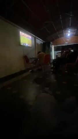 The video shows an interior view of a dimly lit outdoor seating area. A television screen on the left wall displays a sports broadcast, possibly soccer, with colorful graphics. Several people are seated in chairs and on the floor, facing the screen. The seating area appears to be covered by a sloped roof structure. The overall ambiance is casual and social, with a focus on watching the televised event.