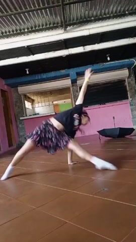 A young female, with purple hair, wearing a black crop top, a black and pink patterned skirt, and white socks, performs a dance routine. She begins in a full split, with one arm extended upwards. She then transitions to a floor move, going from her side to a crawling position, with her body arched. She continues with a series of quick, dynamic movements, including a spin and a bridge-like pose. The dance takes place indoors on a tiled floor, with a pink and blue wall in the background. An open black umbrella is visible on the floor to the right.