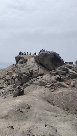 The video shows a view from a rocky hilltop overlooking a city.  Initially, a group of people and bicycles are visible on the rocky hilltop. A person in a red jacket is visible standing near a bicycle on a lower level of the rocks. The camera then pans to show a wide view of a densely built city extending into the distance, partially obscured by a hazy sky. The camera continues to pan slowly across the cityscape, showcasing the urban environment from the elevated vantage point. The ground in the foreground is rocky and sandy.