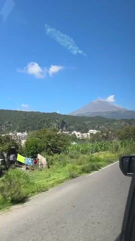 A road runs along the right side of the frame, with a car's side mirror visible. To the left of the road is a grassy verge, followed by lush green vegetation and trees. In the middle ground, a small village with white buildings is nestled amongst the trees. Beyond the village, a large, imposing volcano dominates the background, its peak shrouded in a faint plume of smoke against a clear blue sky with scattered white clouds. The image appears to be taken from a moving vehicle.