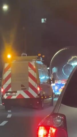The video captures a night scene on a street in Winterthur, Switzerland, showing several vehicles in what appears to be slow-moving or stopped traffic. In the foreground, the rear of a silver car is visible, with its red taillights illuminated. Ahead of it, to the left, is a white utility or road work van, identified by its prominent red and white diagonal striped markings on the rear doors and amber flashing lights on its roof.

The most striking element in the scene is a colossal, dark, circular object being transported on a flatbed truck. This object, possibly a large industrial dish, tank end, or component of a turbine, dominates the mid-ground. Its reflective surface gleams under the ambient light. The large object appears to be wider than a standard lane, suggesting it is an oversized load. A person can be briefly seen near the base of this large object as the video progresses.

The overall illumination is low, characteristic of nighttime, with light sources primarily from vehicle headlights and taillights, flashing amber warning lights, and distant streetlights or building lights. No specific weather conditions are discernible, but reflections suggest a dry surface. There is no legible text visible on any of the vehicles or in the background.