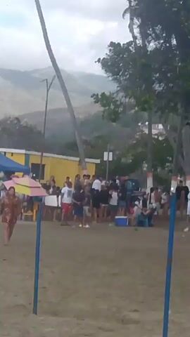 A woman in a black swimsuit is on a sandy beach, holding a leash attached to a small dog. She appears to be running or moving quickly. In the background, a crowd of people are gathered, some sitting and others standing. Palm trees and a hill are visible in the distance. The scene appears to be during the day.