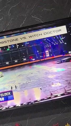 This is a short, shaky video filmed from an angle looking at a television screen. The screen displays a robot combat competition. The words "TOMBSTONE VS. WITCH DOCTOR" are visible at the top of the screen. A digital timer in the lower left corner shows "00:57", then "00:56", "00:55", and finally "00:54". Two robots are visible in the arena. One, identified as Tombstone, is a horizontal spinning disc robot. The other, Witch Doctor, is a vertical spinning disc robot. The robots engage in combat, with sparks flying from the impact. The arena appears to be a metallic enclosure with bright lighting.