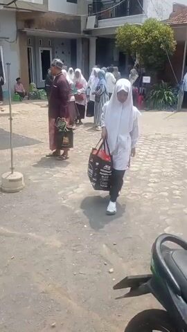 A group of people, mostly women wearing hijabs, are gathered outdoors in what appears to be a courtyard. A young girl in a white hijab and black skirt walks towards the camera, carrying two large shopping bags, one black and one red. In the background, other individuals are walking and standing, some holding bags or other items. The ground is paved with a mix of asphalt and cobblestones. A motorcycle is partially visible in the foreground. The scene suggests an event or gathering is taking place.