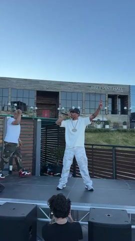 The video shows two men performing on a stage outdoors. The man on the right is singing into a microphone, wearing a white t-shirt, white pants, a baseball cap, and a large gold chain. He is pointing his right hand in the air. The man on the left is wearing a white t-shirt, camouflage pants, and sunglasses. He is holding a microphone in his right hand and gesturing with his left.

Behind them is a building with the text "LISI EVENT HALL" clearly visible. The building has large glass windows and a modern architectural design. There are some potted plants and a wooden fence in front of the building. The sky is clear and blue, suggesting it is daytime, possibly late afternoon or early evening due to the lighting. The overall scene appears to be a performance or concert event. The focus is on the two performers, and the background provides context for the location.
