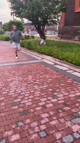 A man with dark hair and a beard, wearing a green long-sleeved shirt and white shorts, runs along a brick pathway. He is wearing bright green athletic shoes. The pathway is wet, with some areas appearing to have collected water. Green grass and a tree are visible to the left of the pathway. A dark-colored building with an orange trim is to the right of the pathway. The sky appears overcast.