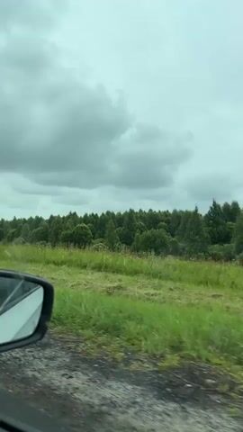 The video is captured from inside a moving vehicle, showing a view through the side window and the side-view mirror. The landscape outside is green and rural, with grassy fields and trees. The sky is overcast with heavy, gray clouds. The side-view mirror reflects a portion of the car's interior and the driver's arm. The camera appears to be looking slightly forward and to the side as the vehicle travels along a road. There are no people clearly visible outside the vehicle. The road surface in the foreground is rough and appears to be gravel or dirt. The overall mood of the scene is somber due to the cloudy weather.