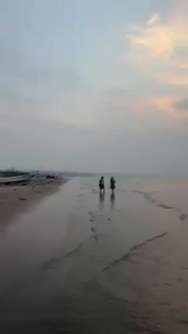 The video shows a beach scene under a sky with a gradient of grey and pink clouds. The wet sand reflects the sky, creating a mirror-like effect. Two children, silhouetted against the light, are walking along the water's edge. To the left, several small boats rest on the sand near other beachgoers. Gentle waves lap against the shore.