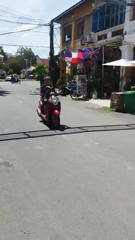 A woman is riding a red scooter down a paved street. She is wearing a dark shirt and light-colored pants. A child is sitting in front of her on the scooter. The scooter is casting a shadow on the street. In the background, there are buildings with awnings and plants. A white car is parked on the right side of the street. The sky is blue and there are some clouds. The video is shot from a slightly elevated perspective, following the scooter.
