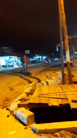 The video depicts an outdoor nighttime scene, possibly a construction site or a roadside area. A makeshift wooden platform is visible in the foreground, surrounded by dirt and concrete blocks. A utility pole stands near the platform. In the background, there's a building with blue accents and signs, and an open-air structure with tables and chairs illuminated by blue lights. Two people are visible standing in the distance near the building. The overall impression is one of an under-construction or temporary outdoor area in a dimly lit environment.