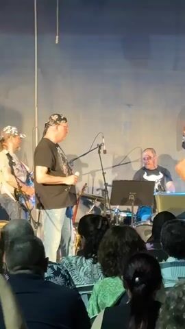 A band is performing on a stage in front of an audience. The band consists of at least five members. On the far left, partially visible, a person wearing a bandana and glasses is holding a microphone and singing. Next to them, a drummer is seated behind a drum kit. In the center of the stage, a man wearing a top hat and a white shirt is holding a microphone and singing. To his right, a man with a beard is playing a bass guitar. Further to the right, a man with glasses is sitting and playing an acoustic guitar. The stage backdrop is a dark blue, textured wall. An object resembling a decorative bottle is hanging from the ceiling. The audience is visible in the foreground, seated and looking towards the stage. The lighting is dim, with spotlights on the performers.