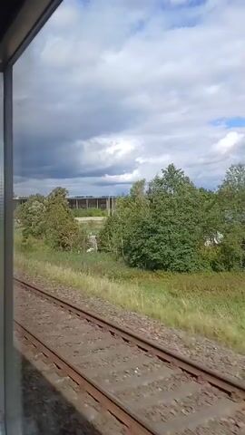 A train is traveling along a track. The view is from inside the train, looking out the window to the right. The sky is cloudy with patches of blue. To the right of the tracks, there is tall green grass and trees. In the distance, there is a long, dark structure, possibly a building or a bridge, with several vertical supports. There are also some telephone poles visible. The train tracks are made of gravel and have parallel metal rails. The video captures the movement of the train as it passes through this rural landscape.