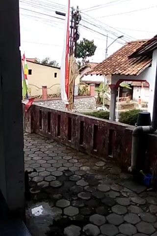 The video shows a view from a covered walkway looking out towards a street and houses. The walkway has a tiled floor and a low red wall. A flag pole with an Indonesian flag is visible. There are also houses with red tile roofs and some trees. The weather appears to be overcast. There are no people visible in the video.