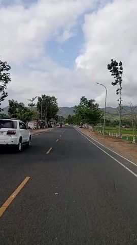 The video shows a view from a moving vehicle on a paved road. On the left side of the road, there is a white SUV. The road is lined with trees and streetlights. To the right of the road, there are rice fields and mountains in the distance. There is also a sign visible that says "POM MINI". The sky is cloudy.