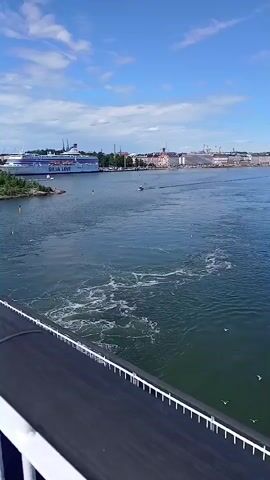 The video shows a wide-angle view of a body of water with a large ferry in the background. The ferry is white with blue and red accents and has the text "BLUE LINE" visible on its side. The water is choppy, with whitecaps and a visible wake behind the ferry, suggesting it is moving. The sky is blue with scattered white clouds. In the foreground, a white railing with a black top rail is visible, implying the video is shot from the deck of another vessel. The deck itself appears to be painted blue and is wet in some areas, reflecting the sky and clouds. The scene is calm and scenic, suggesting a pleasant day for a boat trip.