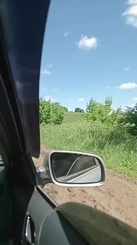 The video shows a view from inside a car driving on a dirt road. The car is moving forward, and the camera is looking out the passenger side window and through the windshield. The road is muddy and bumpy, with green grass and trees on either side. The sky is blue with a few white clouds. The side mirror of the car is visible, reflecting the road and the surrounding scenery. The interior of the car, including the dashboard and door panel, is also visible. The scene suggests a rural or off-road driving experience on a sunny day.