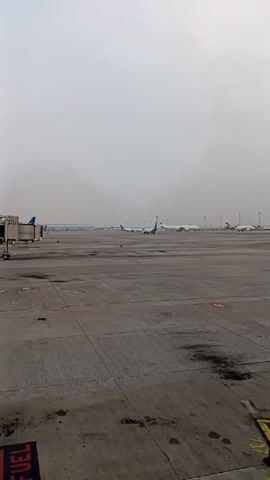 The video captures an airport tarmac on a cloudy day. Several airplanes are visible parked on the tarmac, some with visible airline branding. The tarmac itself is concrete with some wet patches and markings. In the foreground, there are traffic cones and yellow lines indicating boundaries. The sky is overcast, contributing to a muted color palette. No people are clearly visible or interacting in the scene, and the focus is on the static environment of the airport.