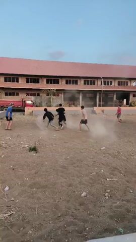 A group of children is playing soccer in a dusty, outdoor area. There is a large building with a red roof in the background, and a red truck parked to the left. The children are running and kicking a soccer ball, creating dust clouds as they play. One child in a blue shirt appears to be a goalie. The sky is clear and blue.