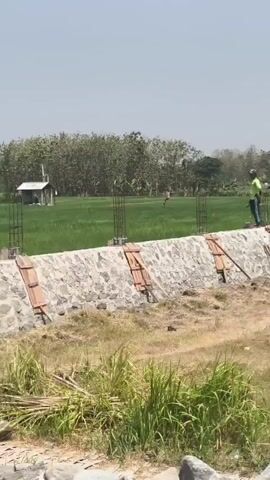 The video shows an outdoor construction scene with several people working. The environment appears to be agricultural with green fields and trees in the background. In the foreground, there is a stone wall being constructed, with wooden supports and rebar visible. Workers are seen near the wall, some appearing to be directing or supervising. Further back, there are temporary shelters and piles of construction materials like sand and gravel. The weather seems to be sunny.