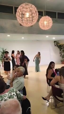 A video shows a group of people at an indoor event, possibly a wedding reception or party. Two large, spherical light fixtures hang from the ceiling. In the background, several women in formal dresses are dancing or being led in a dance by a man in a casual outfit. On the right side of the frame, several women are seated at tables, watching the activity. In the foreground, people are seated at tables, with one person in a light-colored shirt looking towards the dancers. The environment appears to be a banquet hall with a light-colored floor and white walls, decorated with plants.