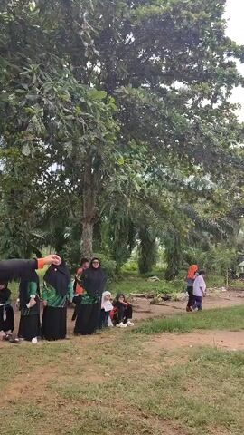 A group of people are gathered outdoors in a grassy area with trees. In the foreground, a person wearing an orange shirt and black pants is facing away from the camera, with their right arm raised. To their left, a group of women wearing headscarves and long dresses stand together. Several children are also present. In the background, two people are interacting near a building, and another person is standing alone. The environment appears to be a park or a recreational area.