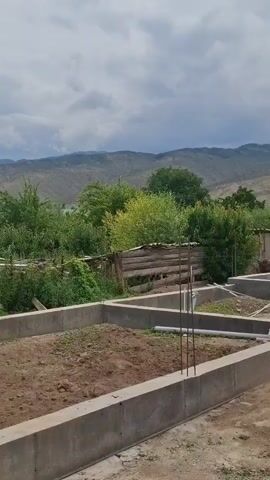 The video shows a construction site with a concrete foundation laid out for a building. The foundation is rectangular and appears to be partially filled with dirt. In the background, there are trees and mountains under a cloudy sky. A wooden fence is visible behind the foundation, and some metal rods are sticking out of the concrete. The overall scene depicts an unfinished construction project in a rural or semi-rural environment.