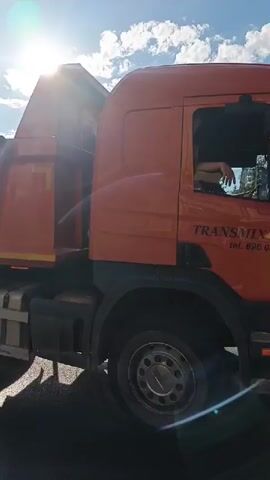 A dump truck is parked on the side of a road. A trailer is attached to the back of the dump truck. The trailer is carrying a road roller. The sun is shining brightly in the sky, and there are some clouds. The road roller is orange and has the brand name "HAMM" visible on it. The trailer has ramps that are folded up. There is a white car in the background.