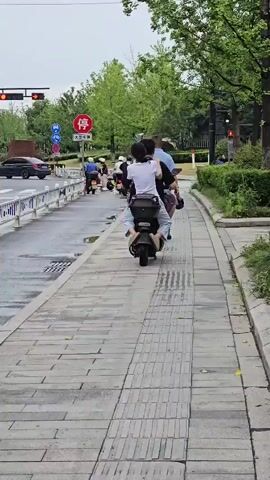 A group of people on electric scooters are seen moving along a street and sidewalk. The weather appears to be overcast. On the street, a truck is visible, and several electric scooters are lined up at a red traffic light. On the sidewalk, two individuals are riding on one scooter, with one person sitting behind the other. Another person is walking on the sidewalk. The environment includes trees, streetlights, and traffic signals. The sidewalk is paved with concrete tiles and features a tactile paving strip.